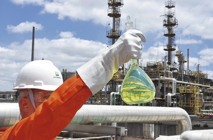 Trabalhador da Petrobras posicionado em uma refinaria de petróleo, vestindo um uniforme laranja, capacete e luva e segurando um frasco que contém uma substância amarelada.