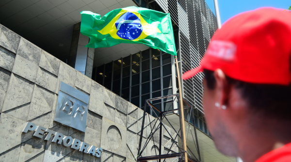 Foto de um homem olhando para o logo da Petrobras afixado na fachada da sede, e, à direita da imagem, a bandeira do Brasil. Crédito: Tânia Rêgo/Agência Brasil.