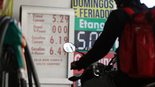 Imagem de um posto de combustível em que, ao fundo, aparecem duas tabelas de preços; à direita, um motociclista esperando para abastecer a moto; e, à esquerda, as bombas de combustível.